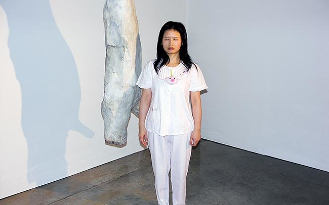 Chun Hua Catherine Dong wears pyjama and walks around in a gallery, posting herself beside art objects with her eyes close