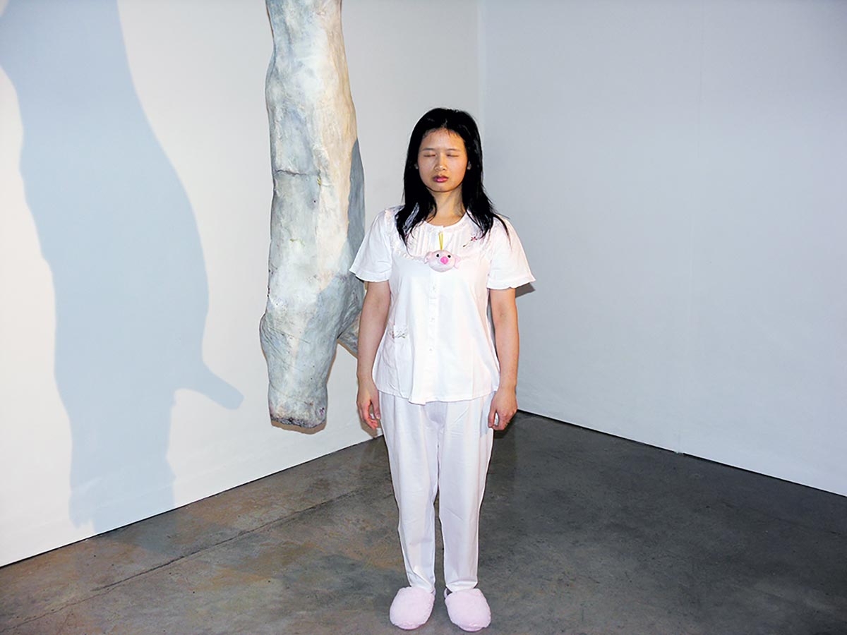 Chun Hua Catherine Dong  wears pyjama and walks around in a gallery, posting herself beside art objects with her eyes close