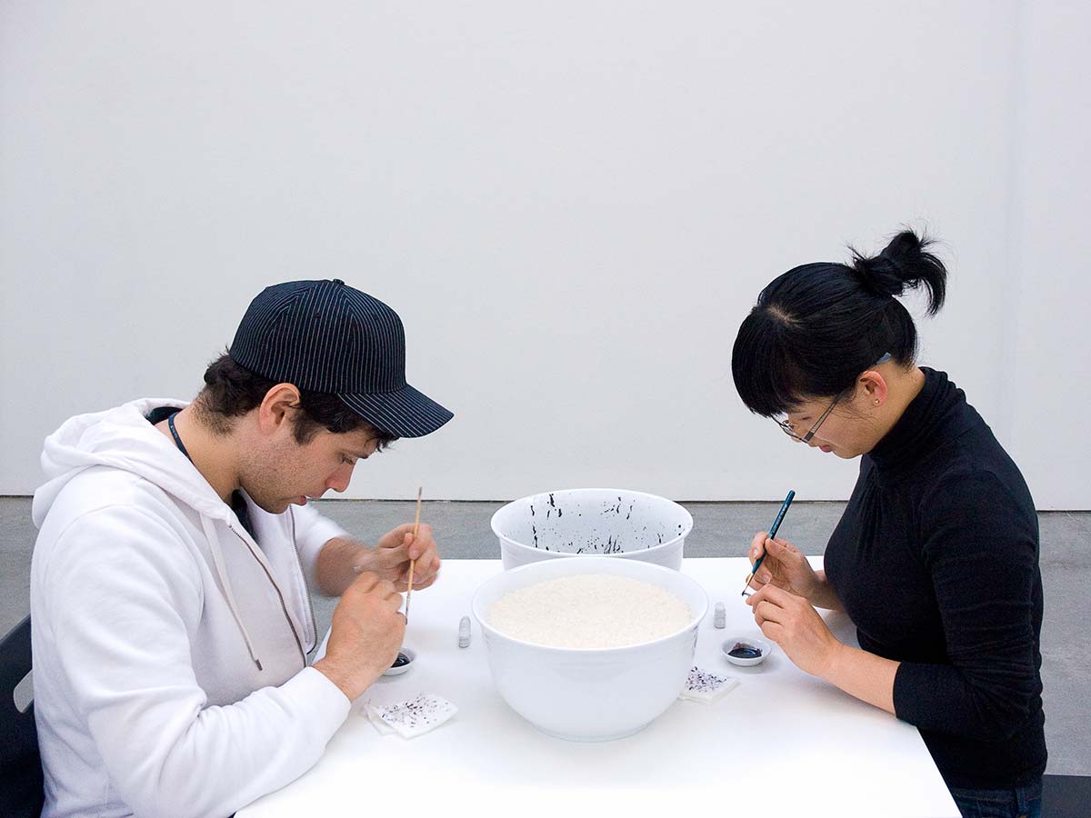Chun Hua Catherine Dong paints rice with black ink one after one and invites audiences to sit in front of her and paint the rice with her