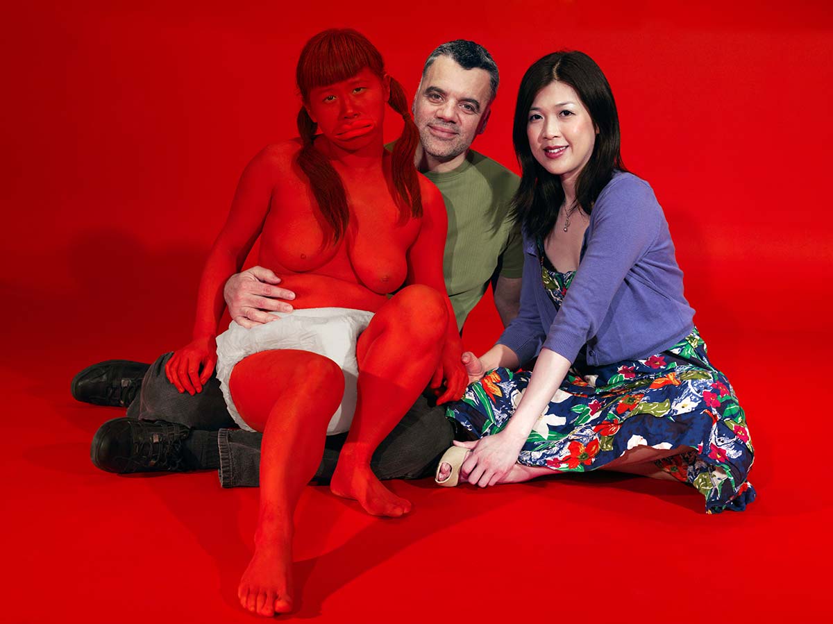 Chun Hua Catherine Dong painted her body red and wore diaper, living with strangers hired from Craigslist in a red room as their child. The red baby is sitting on her father's lap like a giant unhappy baby