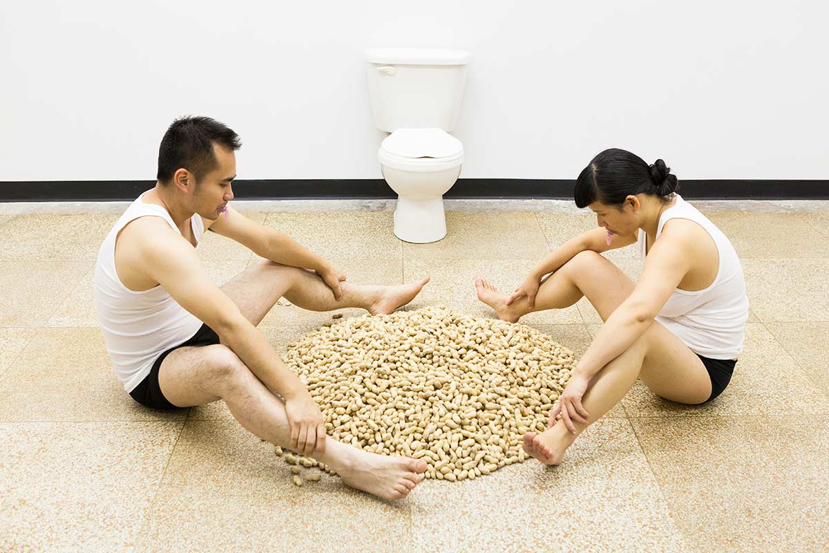 Chun Hua Catherine Dong and her performance partner sit in front of a toilet and look at a big pile of peanuts