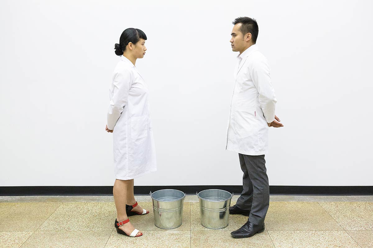 Chun Hua Catherine Dong and her performance partner wear lab coats, standing in front of two buckets and looking at each other