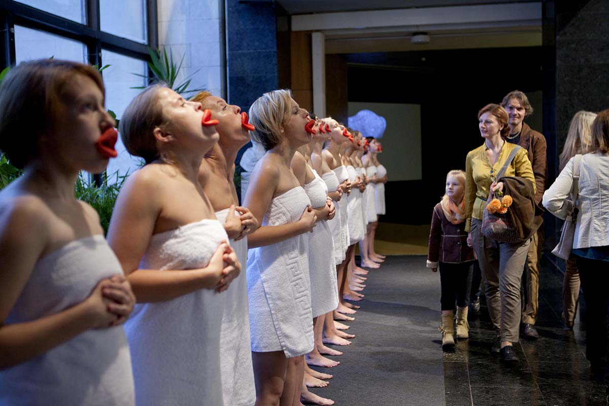 Chun Hua Catherine Dong's performance at Kaunas Biennial: nineteen girls wear red mouthpieces and white bath towels, standing in a row, repeating three still gestures: standing, kneeing, and lying on the floor.