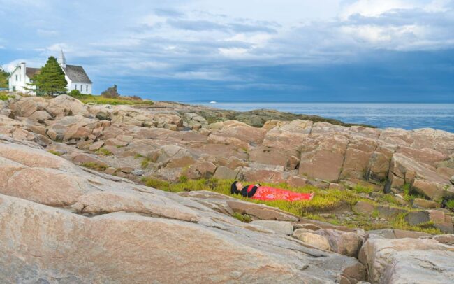 a girl is covered by a red blanket and lies beside water on rocks