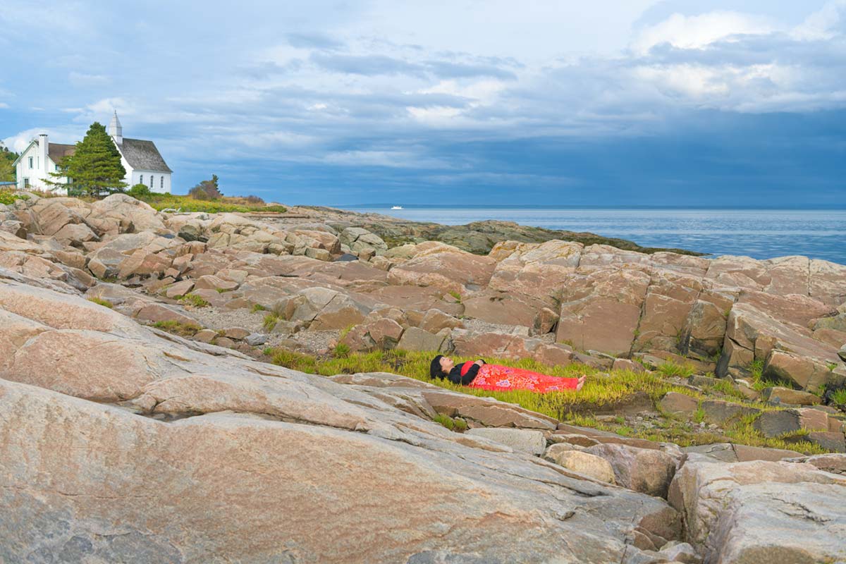 a girl is covered by a red blanket and lies beside water on rocks