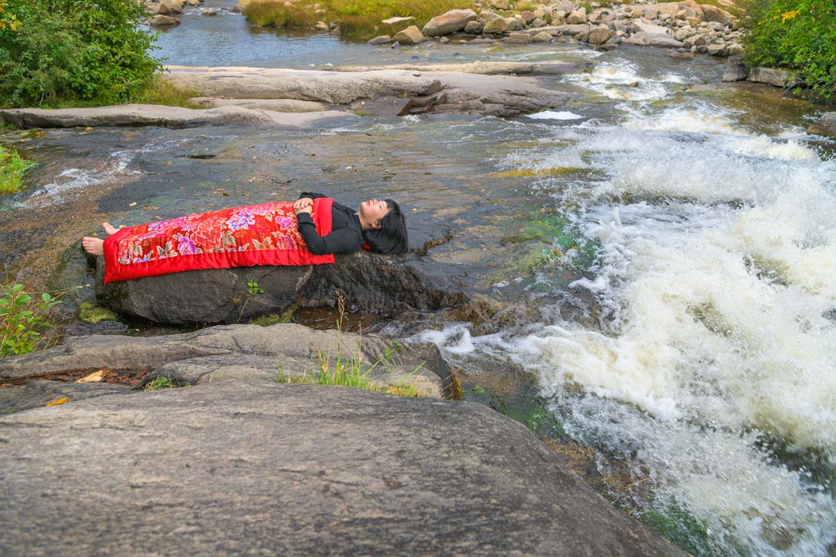 a girl is covered by a red blanket and lies beside water