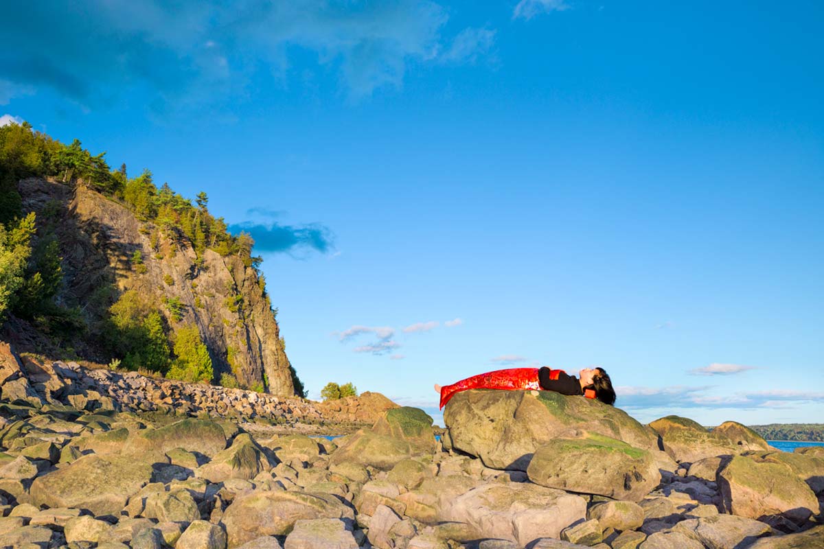 a girl is covered by a red blanket and lies beside Saint Lawrence River on a large rock