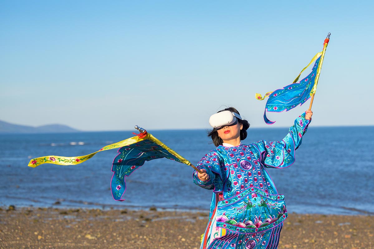 Chun Hua Catherine Dong wears Oculus VR headset and a blue Chinese opera costume with two flags beside The St. Lawrence River.