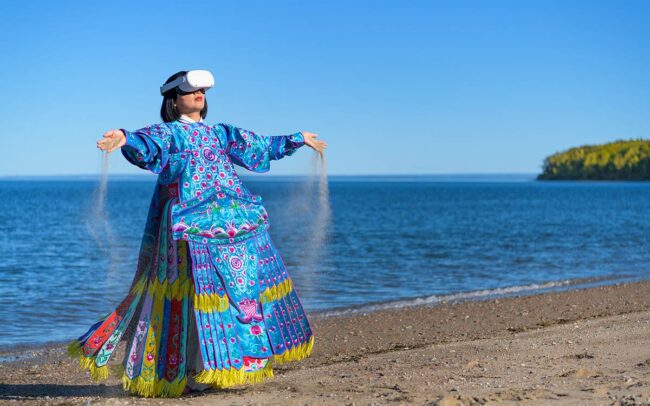 Chun Hua Catherine Dong wears Oculus VR headset and a blue Chinese opera costume with sand on her hands beside The St. Lawrence River.