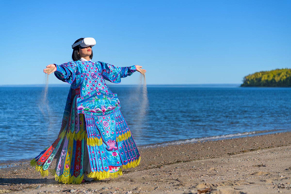 Chun Hua Catherine Dong wears Oculus VR headset and a blue Chinese opera costume with sand on her hands beside The St. Lawrence River. 