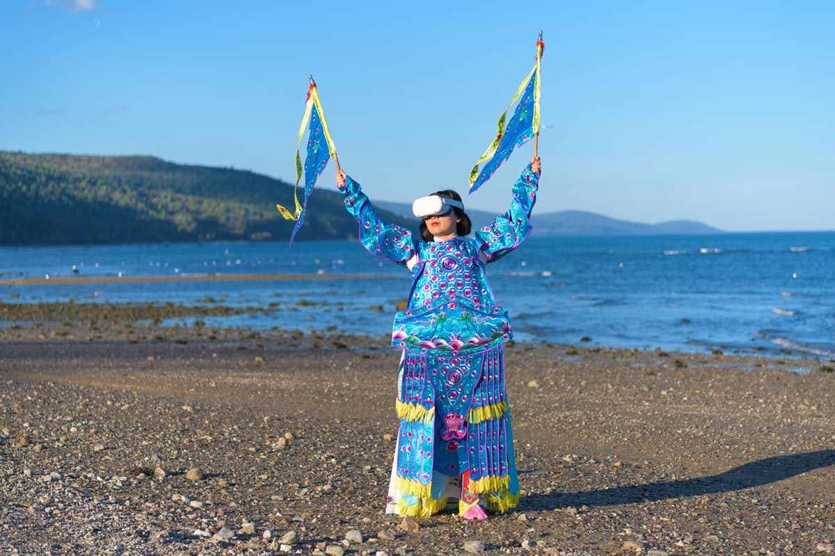 Chun Hua Catherine Dong wears Oculus VR headset and a blue Chinese opera costume with two flags beside the St. Lawrence River.