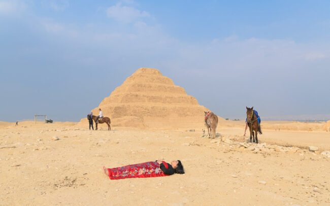 Chun Hua Catherine Dong sleeps in front of the famous pyramid, Dahshur , covered by a red blanket, with a horse, a donkey, Cairo