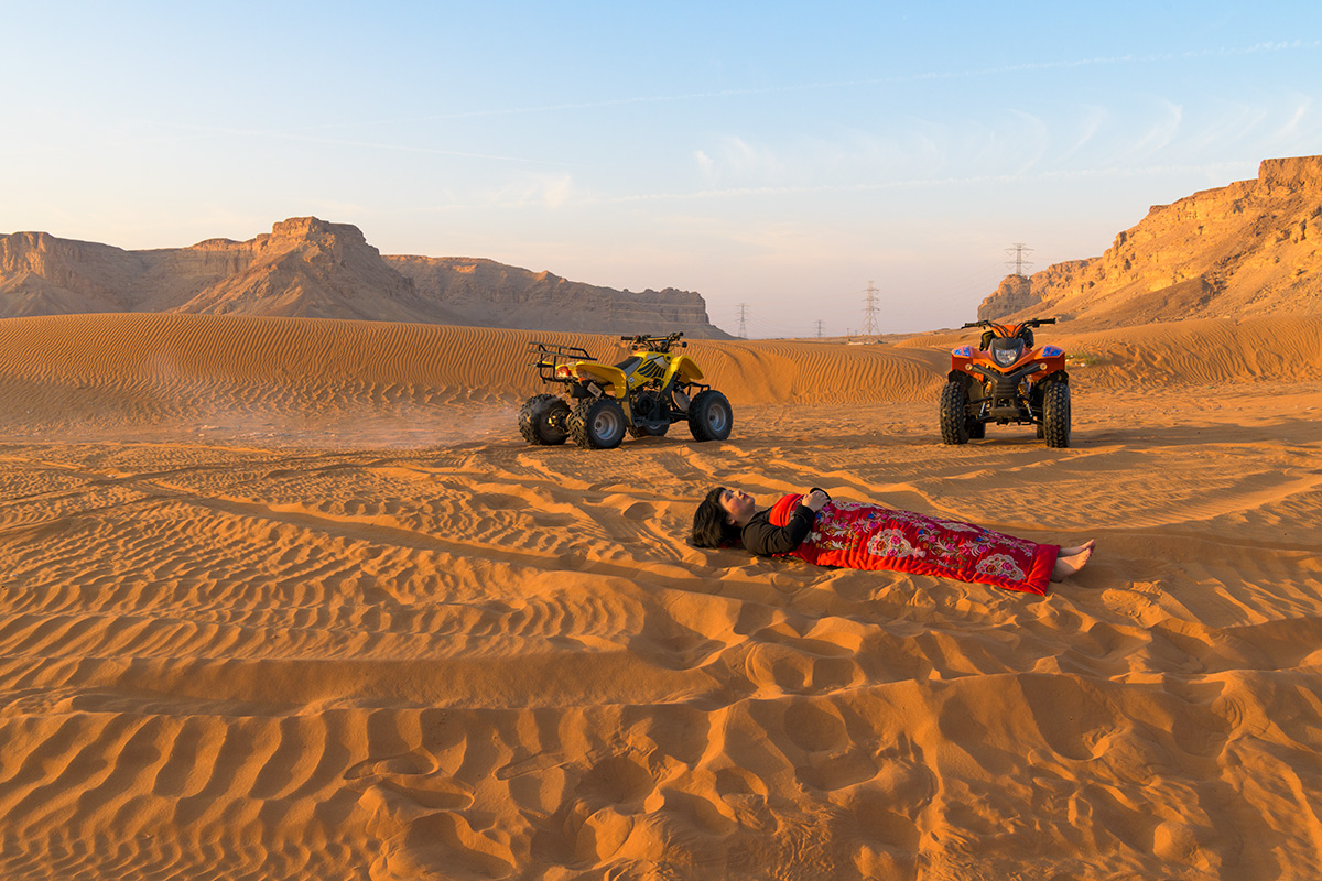 Chun Hua Catherine Dong’s body is covered by a Chinese red blanket, Dong lies in front of two sand bike on Red Sand Dunes in Riyadh, Saudi Arabia.