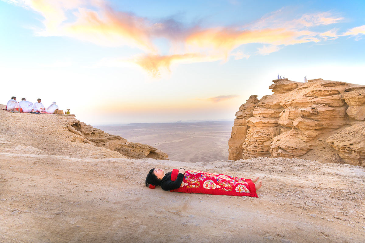 Chun Hua Catherine Dong sleeps at edge of the world, Riyadh, Saudi Arabia, her body is covered with a Chinese red blanket, four Saudi men sit behind her.