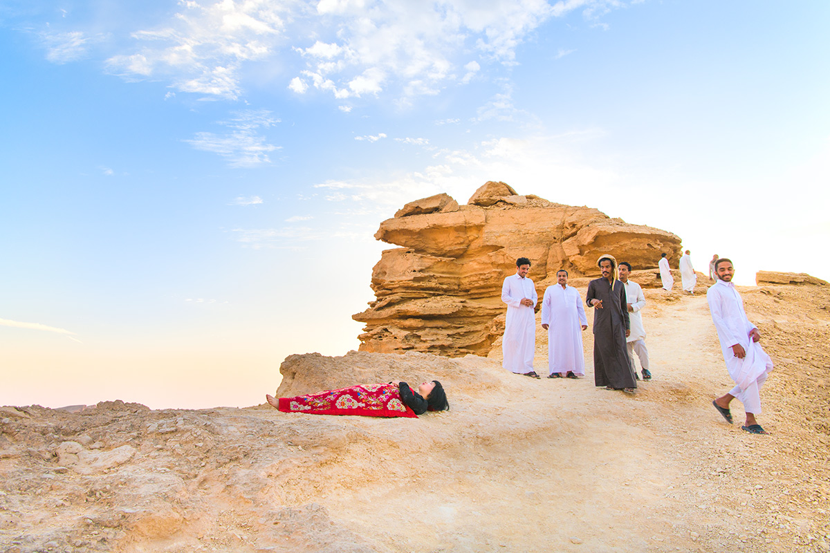 Chun Hua Catherine Dong sleeps at edge of the world, Riyadh, Saudi Arabia, her body is covered with a Chinese red blanket, four young Saudi men look at her.