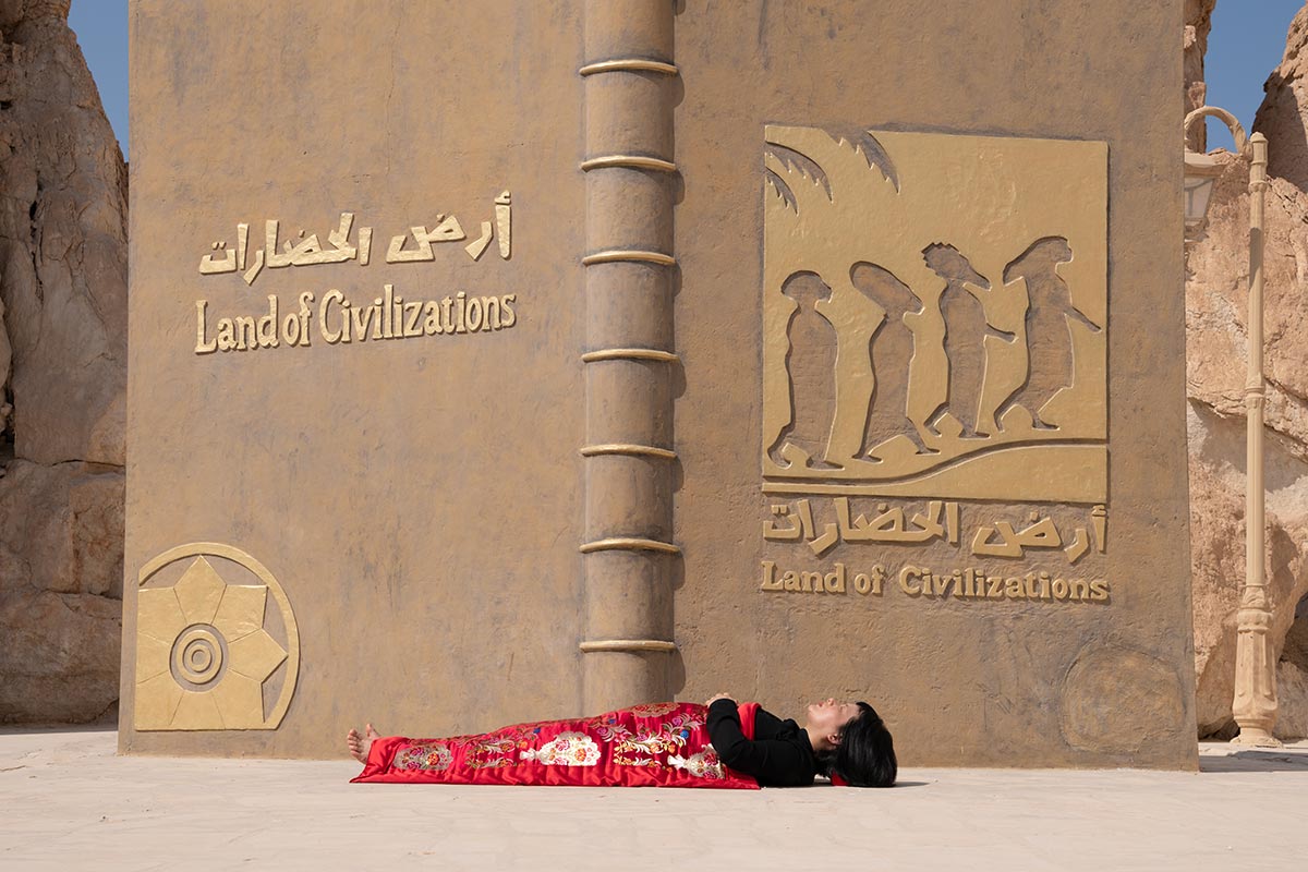 Chun Hua Catherine Dong sleeps in front of a big book called land of civilizations, Al-Qara Hill , Saudi Arabia, her body is covered with a Chinese red blanket.