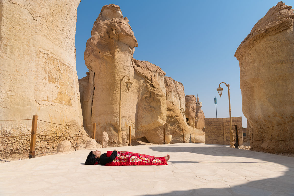 Chun Hua Catherine Dong sleeps in front of group strange rocks, Al-Qara Hill , Saudi Arabia, her body is covered with a Chinese red blanket