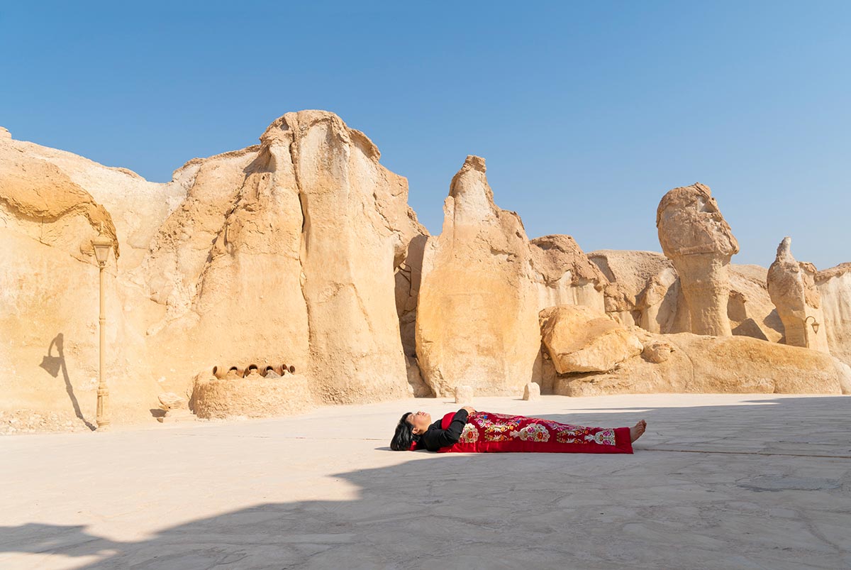 Chun Hua Catherine Dong sleeps in front of group strange rocks, Al-Qara Hill , Saudi Arabia, her body is covered with a Chinese red blanket