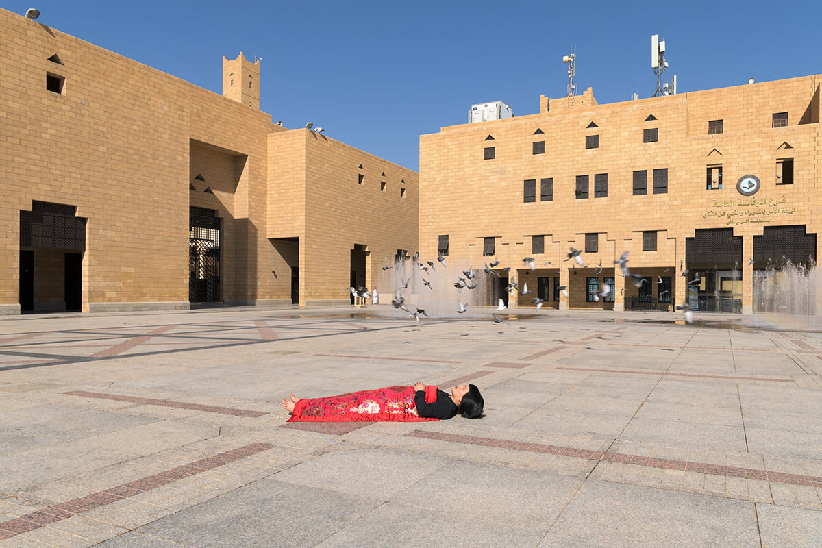 Chun Hua Catherine Dong sleeps in front of Imam turki bin aboullah grand mosque, Riyadh, Saudi Arabia, her body is covered with a Chinese red blanket. Lots of birds fly up behind her