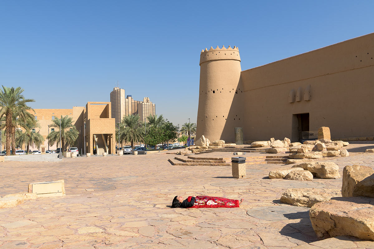 Chun Hua Catherine Dong sleeps in front of Masmak foutress, Riyadh, Saudi Arabia, her body is covered with a Chinese red blanket.