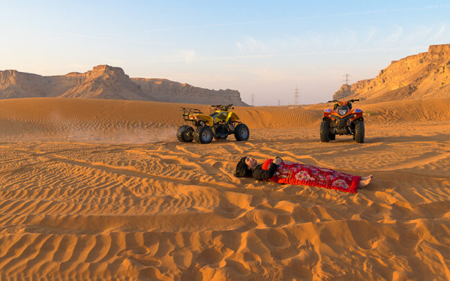 Chun Hua Catherine Dong’s body is covered by a Chinese red blanket, Dong lies in front of two sand bike on Red Sand Dunes in Riyadh, Saudi Arabia.