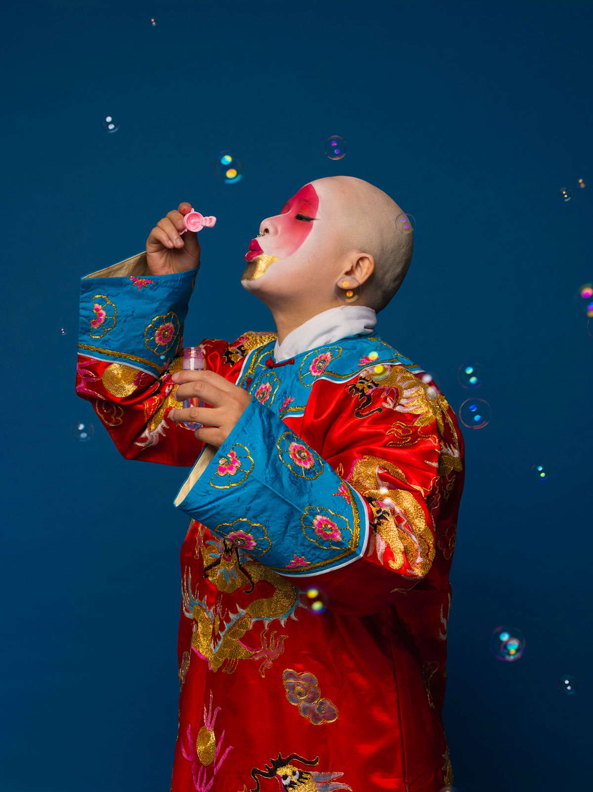 a girl wearing Beijing Opera costume with shaved head and blows bubbles